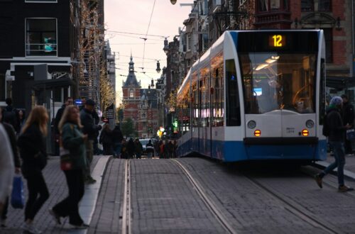 Blå og hvid letbane kører gennem en bygade med kontorbyggeri i dagslys